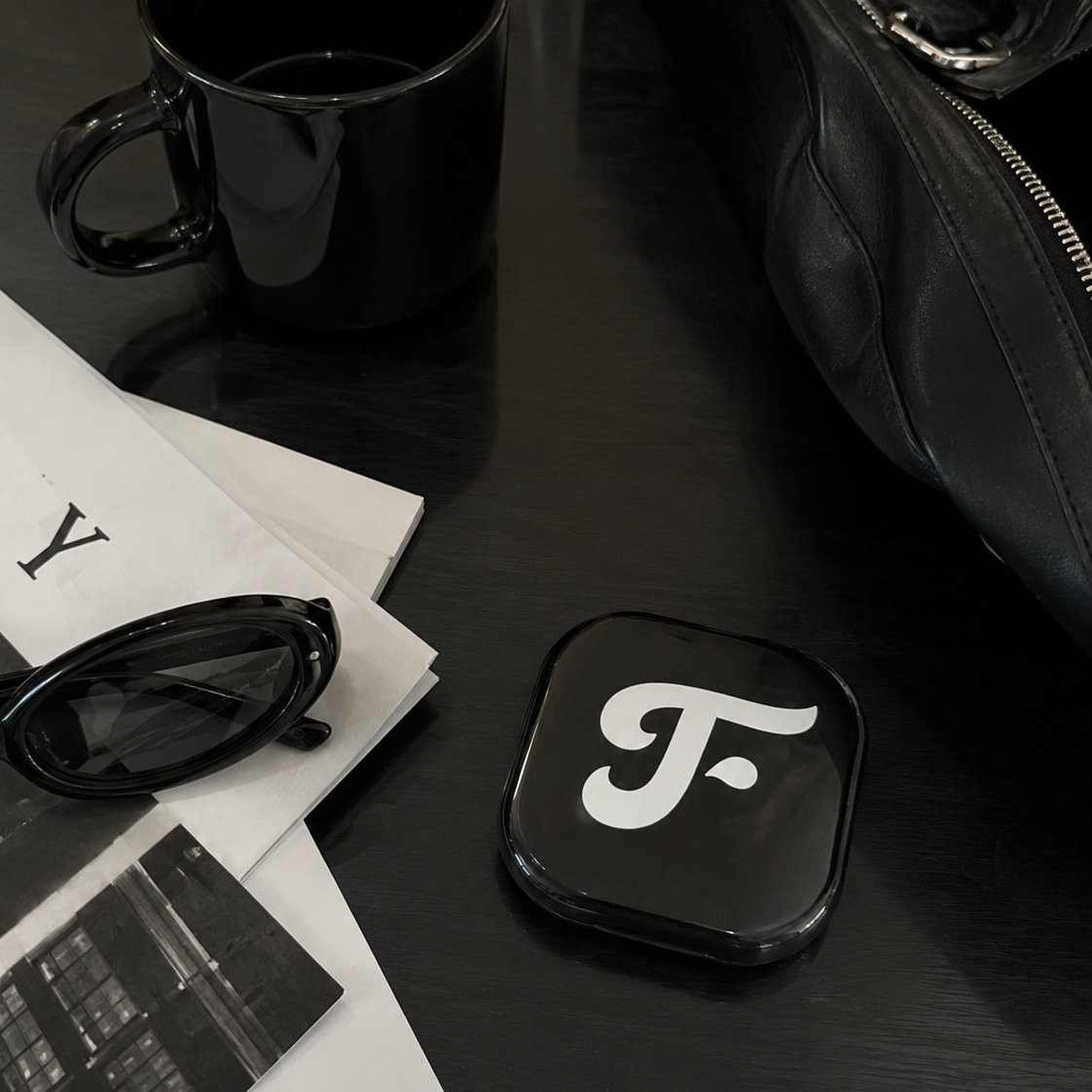 Black coaster with a white logo on a dark surface with sunglasses and papers.