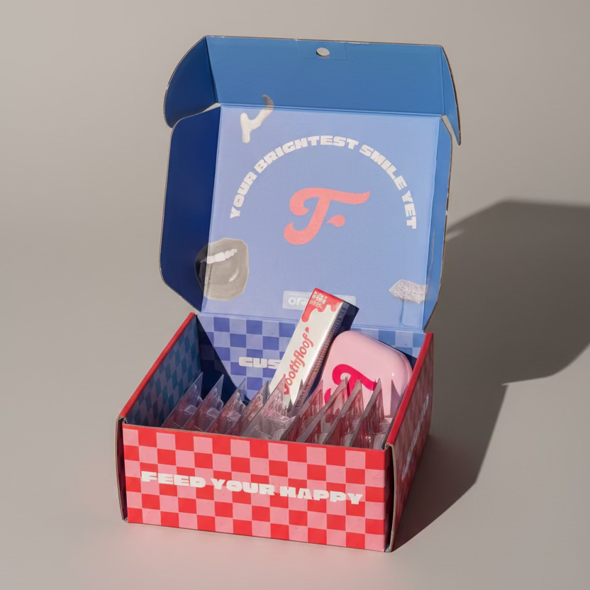 Open box with orawraps and a pink container, featuring a checkered pattern and visible branding.